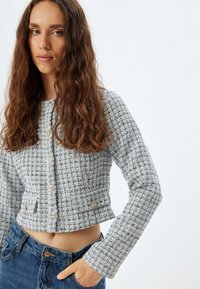 Grå och vit texturerad kort jacka med runda knappar, långa ärmar och två framfickor, parat med högmidjade blå jeans.