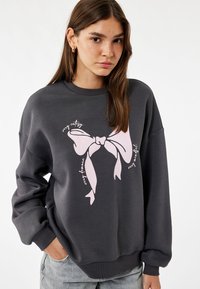 Grå sweatshirt med en stor pink sløjfe grafisk motiv i midten. Har en afslappet pasform, ribbet hals og ærmer. Blød stofstruktur.