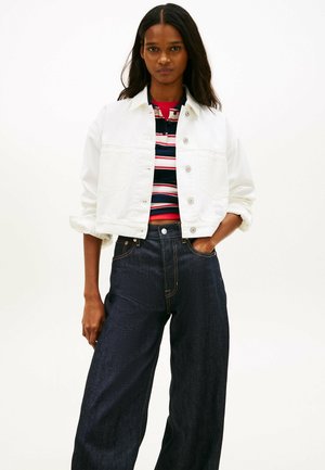 Vrouw met donker haar draagt een wit jack met knopen over een rood-wit-blauw gestreept shirt, gecombineerd met donkere, hooggetailleerde jeans met wijde pijpen.