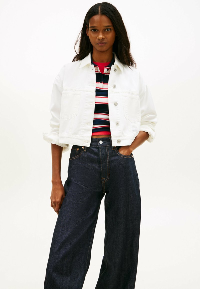 Femme aux cheveux foncés porte une veste blanche boutonnée sur un haut rayé rouge, blanc et bleu marine, associée à un jean large taille haute foncé.