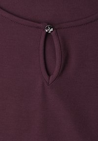 Gros plan sur un tissu bordeaux avec une petite découpe en forme de goutte près de l'encolure, maintenue par un seul bouton argenté.
