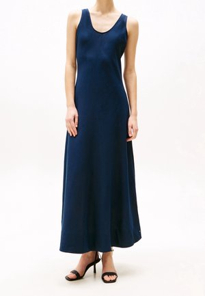 Vrouw in een mouwloze, marineblauwe maxi-jurk met zwarte hoge hakken, staand voor een effen witte achtergrond.