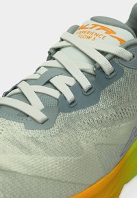 Gros plan sur une chaussure de sport vert clair avec des lacets blancs, une semelle jaune et "Altra Experience Flow 3" imprimé en orange sur la languette.