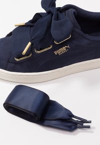 Sneaker Puma Basket bleu marine avec de larges lacets en satin noués en nœud, œillets dorés et une paire supplémentaire de lacets lâches assortis en dessous.