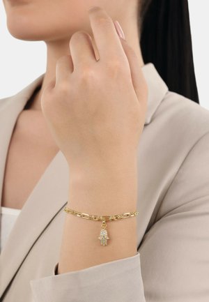 Pulsera de cadena de oro con un charm en forma de mano con intrincados detalles, llevada en una muñeca de piel clara. La pulsera tiene una textura suave.