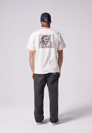 Mand iført hvid T-shirt med lyserød tegneserieglobus og tekst, mørke jeans og marineblå kasket, stående med ryggen til mod ensfarvet baggrund.