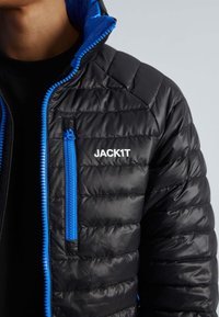 Zwarte pufferjack met blauwe ritsaccenten en een logo. Beschikt over een gewatteerde structuur en een hoge kraag. Ritszak op de borst.