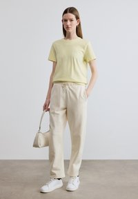 Ανοιχτό κίτρινο t-shirt με κοντό μανίκι, κρεμ παντελόνι με κορδόνι, λευκά αθλητικά παπούτσια. Το μοντέλο κρατάει μία μικρή μπεζ τσάντα χειρός. Ομαλό ύφασμα, άνετη εφαρμογή.
