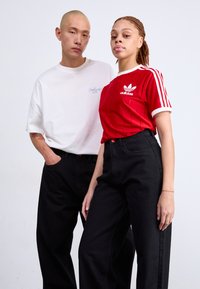 Dva modela poziraju nasuprot jednostavne pozadine. Jedan nosi bijelu pamučnu majicu, a drugi nosi crveni Adidas top s prugama i tri bijele pruge. Oboje nose crne traperice.