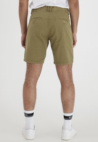 Man wearing olive green dotted shorts, white socks with black stripes, and white sneakers standing facing away on a plain white background.