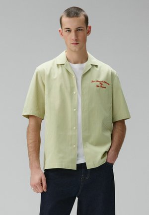 Young man with short hair wears light green vertical striped short-sleeve shirt over white t-shirt and dark pants, standing against plain background.