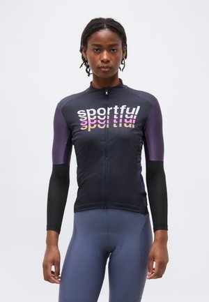 Moteris dėvi aptemptą juodai violetinį dviračio džersį su „sportful“ logotipu ir aptemptus mėlynus dviračio šortus, stovinti priešais vienodą baltą foną.