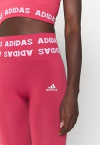 Rosa träningsoutfit bestående av en kort topp och leggings, båda med kontrasterande vit "ADIDAS"-logotyp och band. Mjuk, strukturerad tyg.