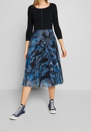 Vrouw draagt een zwarte top met lange mouwen, een blauw-zwart gemarmerde geplooide midi-rok en marineblauwe hoge sneakers en staat tegen een grijze achtergrond.
