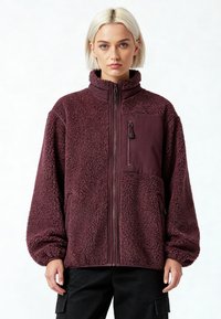 Veste en polaire bordeaux avec un col montant, une fermeture éclair complète et une poche poitrine zippée. Présente une surface texturée et duveteuse ainsi qu'une coupe décontractée.