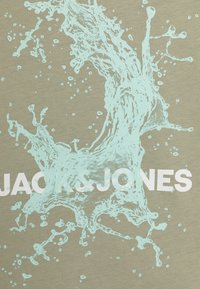 Beiger Stoff mit einem türkisfarbenen Wasserspritzgrafik und kräftigem weißem "JACK & JONES"-Text, der einen dynamischen visuellen Effekt erzeugt.