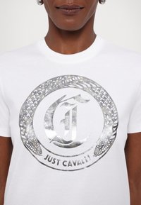 Valkoinen puuvillainen t-paita, jossa on metallinhohtoinen hopeinen logomuotoilu tekstillä "JUST CAVALLI" ja koristeellinen käärmepatterni ympärillä.