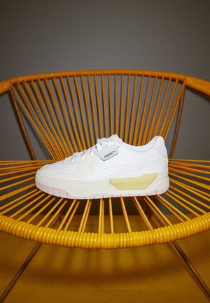 Baskets blanches avec une texture subtile et une semelle en caoutchouc, comportant un accent jaune pâle, un design à lacets et un logo PUMA sur le côté.