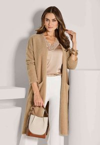 Frau in beigem, flauschigem Cardigan, Oberteil mit Satin und Spitzenrand, weißen Hosen, die eine beige und braune Handtasche hält, steht vor weißen geometrischen Formen.