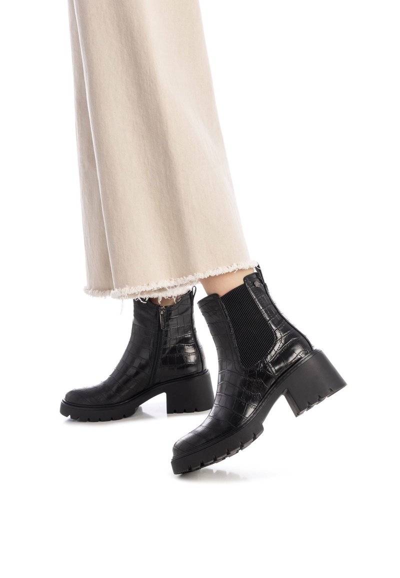 Botas de tobillo de imitación de cuero de cocodrilo negro con tacón grueso, paneles laterales elásticos y suela texturizada; combinadas con pantalones claros y deshilachados.