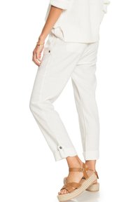 Pantalons en lin blanc avec des ourlets roulés et des détails de boutons, associés à des sandales espadrilles à brides marron. Texture lisse avec une coupe décontractée.