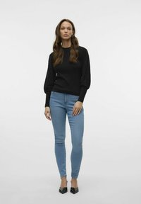 Zwarte trui met gestructureerde ballonmouwen, gecombineerd met lichtblauwe skinny jeans en zwarte puntige hakken. Effen achtergrond.