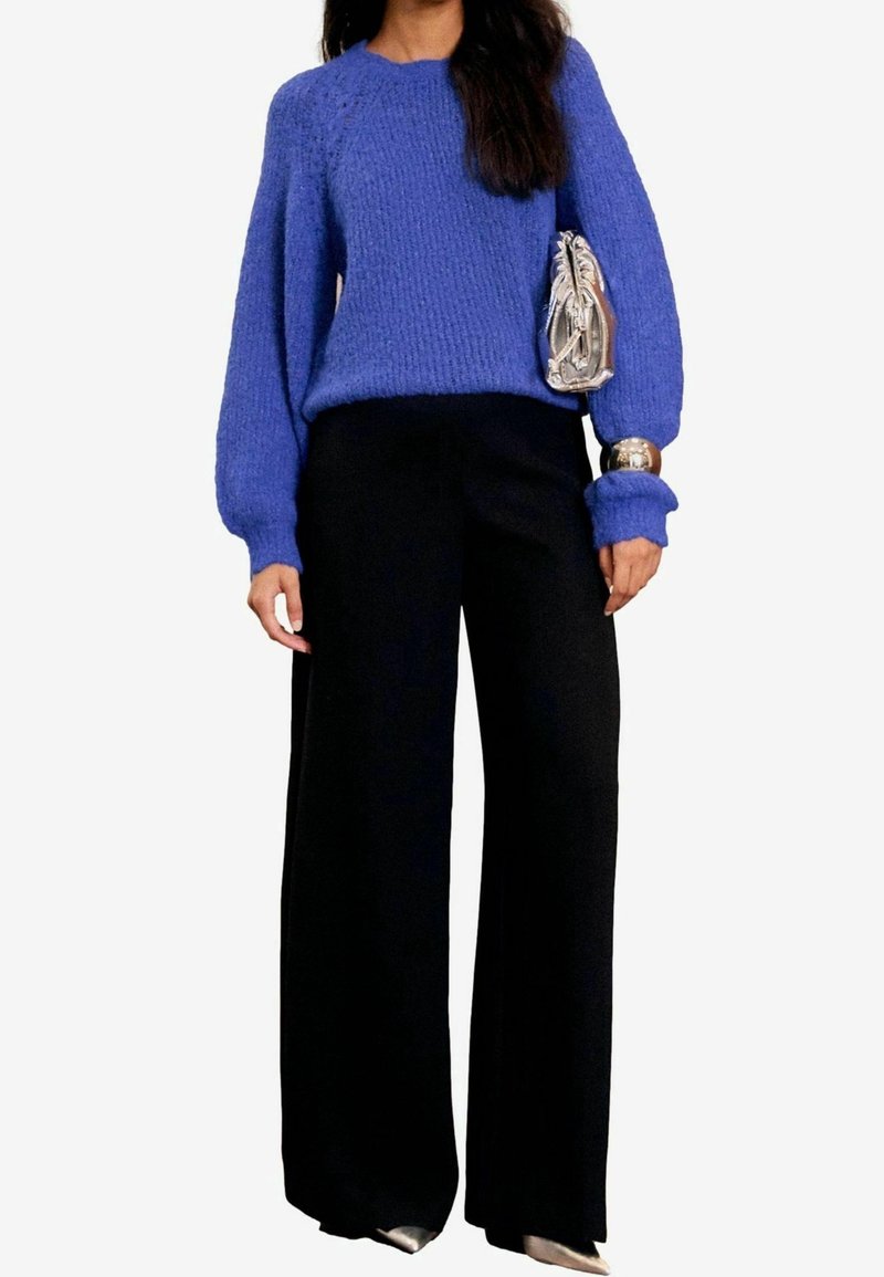 Pull bleu tricoté avec manches longues bouffantes, associé à un pantalon noir à jambes larges. Tient un sac à main argenté texturé.