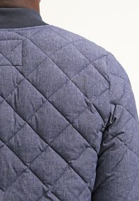 Prošívaná modrá bunda s texturovanou tkaninou a diamantovým vzorem, s žebrovaným límcem. Materiál vypadá měkký a teplý.