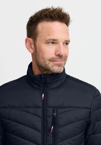 Marineblå pufferjakke med høy krage, med horisontal søm, glidelåslommer og røde og hvite glidelåstrekk. Myk tekstur.