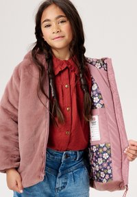 Roze fleece- gevoerde jas met kap, voorzien van een bloemenpatroon aan de binnenvoering, gecombineerd met een rode overhemddas en blauwe denim shorts.