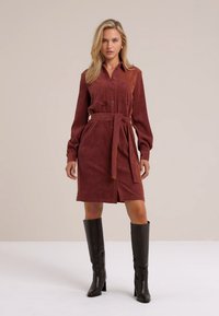 Abito in velluto corduroy color marrone con chiusura a bottoni frontale, maniche lunghe e cintura coordinata, con uno spacco frontale e tasche laterali, abbinato a stivali neri.
