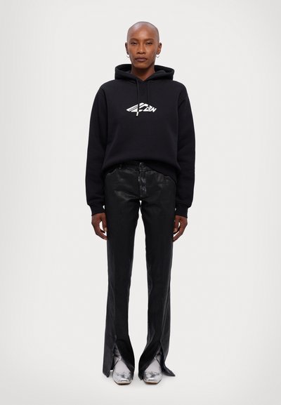 Black sweatshirt with a white logo, paired with shiny black flared pants featuring side slits, complemented by metallic silver shoes.