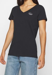 Žena v černém tričku s výstřihem do V značky Tommy Jeans a světlých modrých džínách, zobrazená od ramen po boky.
