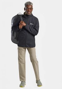 Black zip-up jacket with logo, beige pants, and gray sneakers. Accessories include a gray backpack. Smooth fabric, relaxed fit.