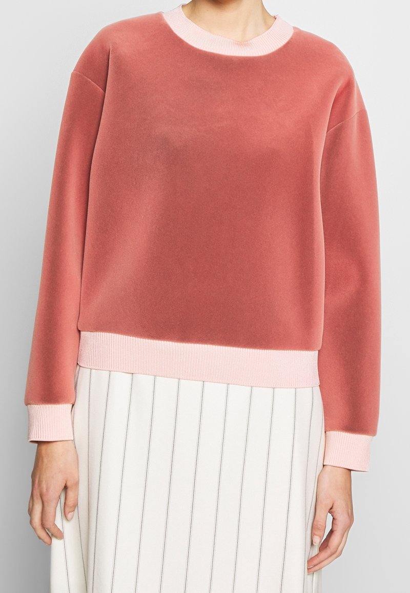 Sudadera rosa con una textura suave y aterciopelada, puños y dobladillo acanalados. Presenta un cuello redondo y un corte relajado, combinada con una falda de rayas.