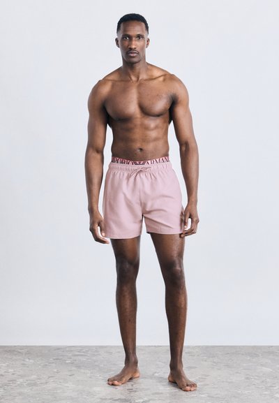 Μυώδης άνδρας όρθιος, ξυπόλητος, φοράει ροζ μαγιό με εμφανές λογότυπο Emporio Armani στη μέση, πάνω από απλό φωτεινό φόντο.