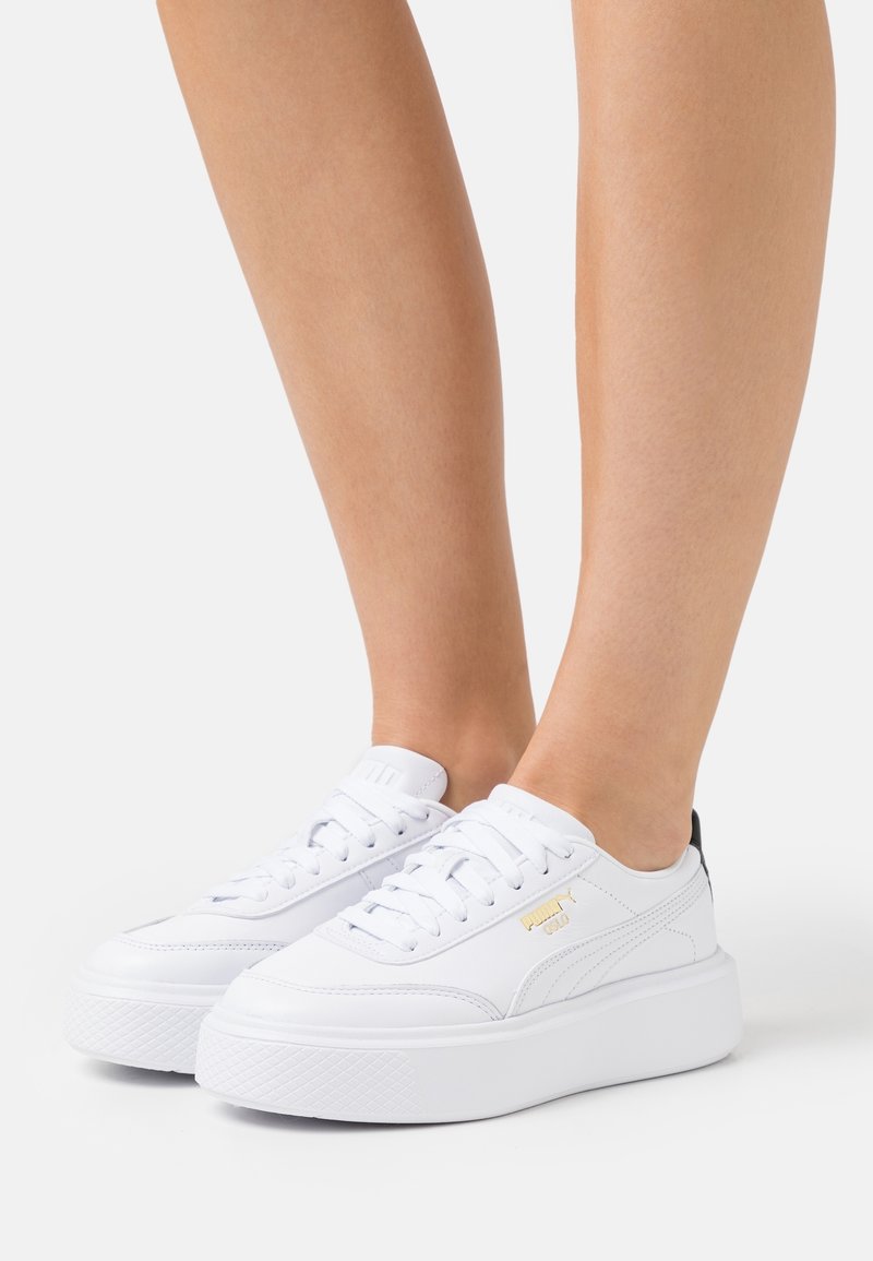 Białe sneakersy na platformie z gładkiej skóry, zaokrąglonym przodem, płaskimi sznurówkami, złotym logo oraz kontrastującym czarnym akcentem na pięcie.