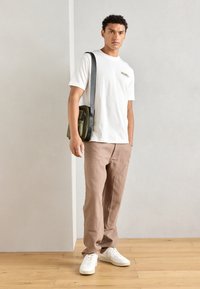 Hvit t-skjorte med lite logo, beige løstsittende bukser, hvite sneakers og en grønn crossover-veske med svart stropp, stående på tregulv.