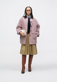 Κόκκινο και λευκό καρό μεγάλο σακάκι με καφέ γιακά, μπλε πουλόβερ, μπεζ πλισέ φούστα και καφέ μπότες μέχρι το γόνατο, κρατώντας ένα μπεζ φάκελο.