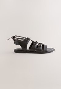 Sandale gladiateur en cuir noir avec plusieurs brides, boucles argentées et fermeture à nouer à la cheville sur semelle plate, vue de côté sur fond neutre.