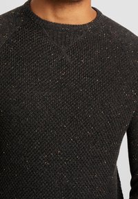 Ciemnoszary sweter dziany z teksturowanym wzorem, z drobnymi złotymi i czerwonymi akcentami. Okrągły dekolt i rękawy raglanowe.
