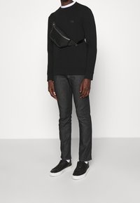 Man som bär en svart långärmad skjorta, mörka jeans, svarta sneakers med vita sulor och en svart axelväska mot en enkel vit bakgrund.