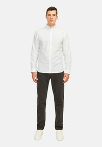 Camisa blanca de manga larga y botones con cuello clásico, combinada con pantalones gris oscuro y zapatillas blancas, de pie frente a un fondo neutro.