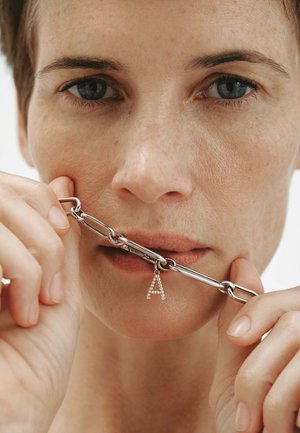 Primer plano de una persona sosteniendo una pulsera de cadena plateada con un colgante en forma de letra A cerca de sus labios, mostrando la textura natural de la piel y cabello corto.