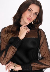 Top nero plissettato con maniche trasparenti a pois, collo alto e dettagli arricciati. Presenta una finitura testurizzata e elementi di design sovrapposti.