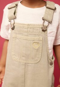 Salopette couleur crème avec bretelles ajustables, quincaillerie en argent et poche avant décorée d'une broderie de smiley. Portée par-dessus un t-shirt blanc.