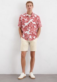 Korte mouw shirt met een rood bloemenpatroon op een witte achtergrond, gecombineerd met beige shorts en witte sneakers met een tan zool.