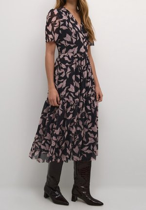 Femme portant une robe midi noire avec un motif de feuilles roses et des bottes noires montantes à talons, debout devant un fond clair uni.