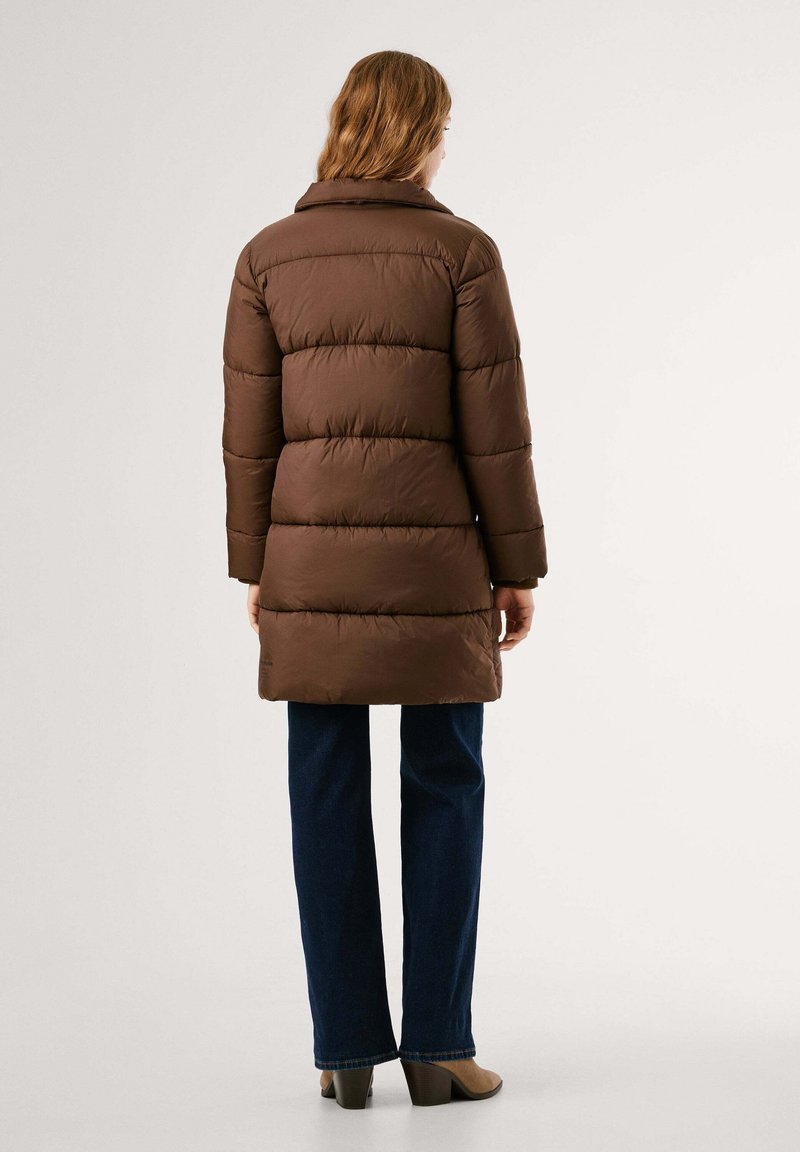 Kobieta ubrana w brązowy ocieplany płaszcz, ciemnoniebieskie dżinsy i beżowe buty na obcasie, stojąca tyłem na jednolitym jasnym tle.