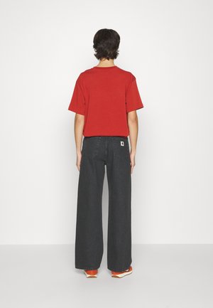 Osoba stojąca tyłem, ubrana w czerwoną koszulkę z krótkim rękawem, czarne dżinsy o szerokich nogawkach oraz pomarańczowo-białe buty na jednolitym tle.